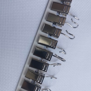 Puede incluir: Un juego de clips de cortina plateados con ganchos. Los clips están dispuestos en una fila sobre un fondo blanco.