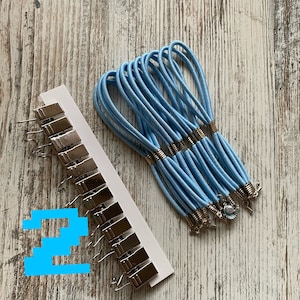 Op de afbeelding: Een set van 15 lichtblauwe elastiekjes voor haar met zilveren metalen accenten en een set van 15 zilveren metalen clips met haken.