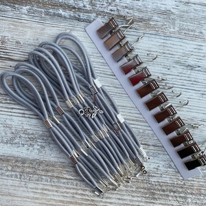 May include: A set of grey elastic hair ties with silver metal accents and a rose charm. The hair ties are arranged in a fan shape and are accompanied by a set of silver metal curtain clips.