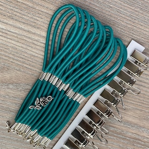 May include: A set of teal elastic loops with silver metal ends and a small rose charm. The loops are bundled together and there are silver metal clips with hooks on a white background.