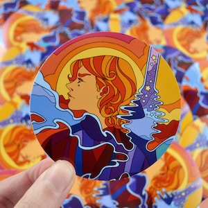 May include: A round sticker with a psychedelic design featuring a person with long red hair, a blue and purple background, and a yellow and orange sunset.