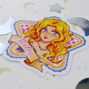 Pu&ograve; includere: Un adesivo con un'illustrazione a cartoni animati di una donna con lunghi capelli biondi ondulati e occhi blu. L'adesivo ha la forma di una stella con uno striscione con la scritta "Haley". Lo sfondo ha un motivo a scacchi rosa e bianchi.