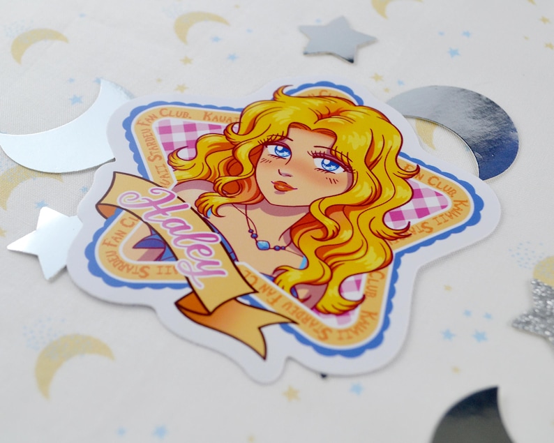 Pu&ograve; includere: Un adesivo con un'illustrazione a cartoni animati di una donna con lunghi capelli biondi ondulati e occhi blu. L'adesivo ha la forma di una stella con uno striscione con la scritta "Haley". Lo sfondo ha un motivo a scacchi rosa e bianchi.
