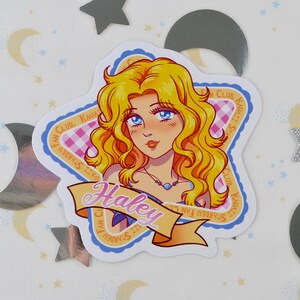 Pu&ograve; includere: Un adesivo con un'illustrazione a cartoni animati di una donna con capelli biondi e occhi blu. L'adesivo ha la forma di una stella e presenta il testo "Haley" su uno striscione. &Egrave; presente anche il testo "Stardew Fan Club".