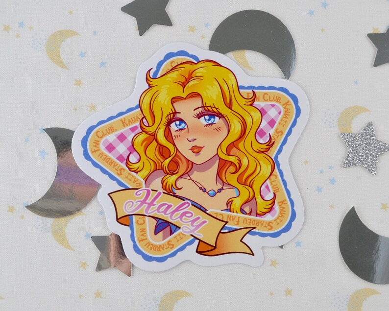Pu&ograve; includere: Un adesivo con un'illustrazione a cartoni animati di una donna con capelli biondi e occhi blu. L'adesivo ha la forma di una stella e presenta il testo "Haley" su uno striscione. &Egrave; presente anche il testo "Stardew Fan Club".