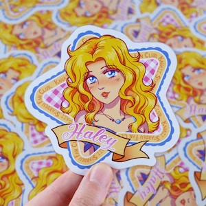 Pu&ograve; includere: Un adesivo con un'illustrazione a cartoni animati di una donna con capelli biondi e occhi blu. L'adesivo ha uno striscione con la scritta "Haley" ed &egrave; circondato da un bordo blu e rosa con la scritta "Stardew Fan Club".