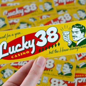May include: A yellow and red Lucky 38 Casino sticker. The sticker features the text "Lucky 38 Casino" and "I went for a spin... but the House always wins!" with an illustration of a man holding a cocktail.