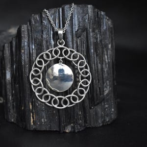 May include: A silver pendant necklace featuring a circular design with a central disc. The pendant hangs from a delicate chain. The necklace is displayed against a dark, textured background, highlighting its metallic shine.