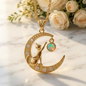 May include: A gold-toned necklace featuring a crescent moon pendant with a cat silhouette reaching toward a round, iridescent opal-like stone. The moon is accented with small, clear, sparkling stones along its curved surface and hangs from a rope chain.