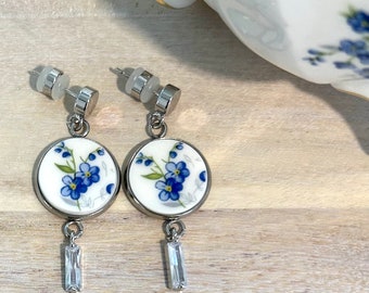May include: A pair of silver dangle earrings with a white porcelain charm featuring blue forget-me-not flowers. Each charm has a clear crystal dangling below.