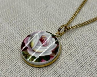 Peut inclure: Un collier doré avec un pendentif rond représentant une rose. La rose a des pétales blancs avec des nuances roses et rouges, et des feuilles vertes. Le pendentif est dans un cadre doré et est suspendu à une chaîne dorée.
