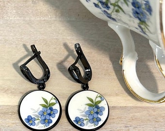May include: A pair of black dangle earrings with white porcelain round charms featuring a blue forget-me-not flower design.