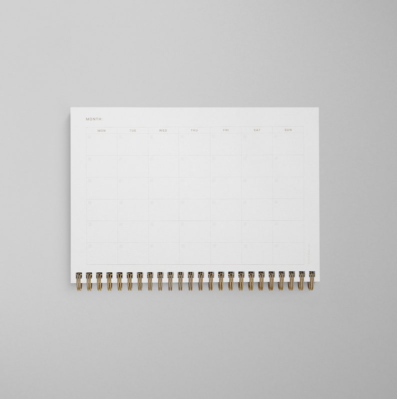 K&ouml;nnte beinhalten: Ein wei&szlig;er Monatskalender mit einer goldenen Spiralbindung. Der Kalender hat ein schlichtes Design mit den Wochentagen, die oben beschriftet sind.