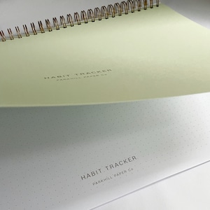 K&ouml;nnte beinhalten: Ein hellgr&uuml;nes Notizbuch mit einer goldenen Spiralbindung. Der Einband hat den Text "HABIT TRACKER PARKHILL PAPER CO." in Schwarz gedruckt. Der hintere Einband ist wei&szlig; mit einem gepunkteten Muster und dem Text "HABIT TRACKER PARKHILL PAPER CO." in Schwarz gedruckt.
