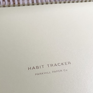 K&ouml;nnte beinhalten: Ein wei&szlig;er Habit Tracker mit einer goldenen Spiralbindung. Der Umschlag tr&auml;gt den Text "HABIT TRACKER" und "PARK HILL PAPER CO" in brauner Farbe.