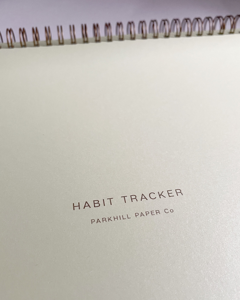 K&ouml;nnte beinhalten: Ein wei&szlig;er Habit Tracker mit einer goldenen Spiralbindung. Der Umschlag tr&auml;gt den Text "HABIT TRACKER" und "PARK HILL PAPER CO" in brauner Farbe.