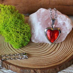 May include: A silver-toned heart-shaped pendant necklace with a vibrant red center. The pendant is displayed on a pink crystal, with a green moss and a wooden slice in the background. The necklace chain is also visible.