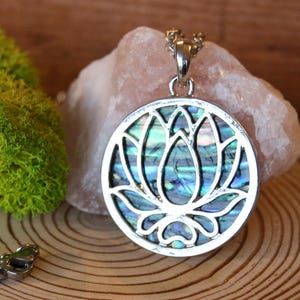 May include: Silver pendant necklace with a circular pendant featuring a lotus flower design. The pendant has a mother-of-pearl inlay with blue and green tones. The necklace is displayed on a wooden slice with green moss and a pink stone.