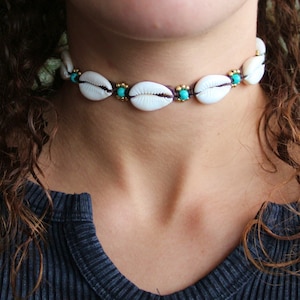 May include: A white cowrie shell choker necklace with gold and turquoise accents. The necklace features multiple white shells, each separated by a gold bead and a turquoise bead. The necklace is worn around the neck.