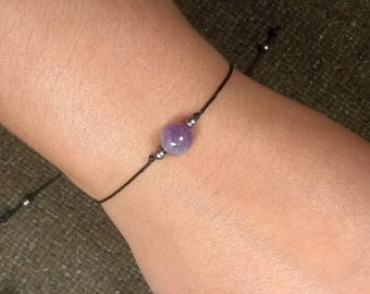Puede incluir: Una pulsera de hilo negro con una cuenta central redonda de piedra preciosa púrpura. La pulsera tiene pequeñas cuentas de color plateado a cada lado de la piedra preciosa. La pulsera está en una muñeca.