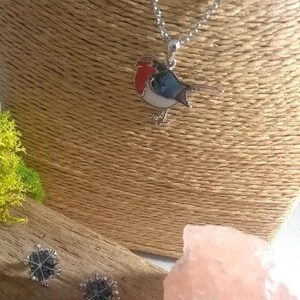 May include: A silver necklace with a bird pendant featuring red, blue, and white enamel. Two snowflake-shaped stud earrings are also visible. The jewelry is displayed on a neutral-toned bust.