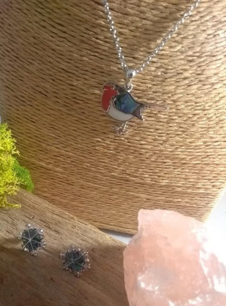 May include: A silver necklace with a bird pendant featuring red, blue, and white enamel. Two snowflake-shaped stud earrings are also visible. The jewelry is displayed on a neutral-toned bust.