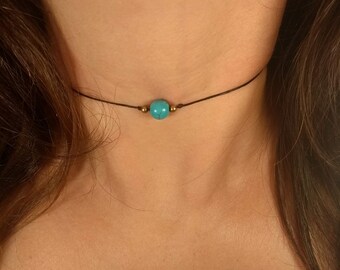May include: A delicate choker necklace featuring a vibrant turquoise bead as the focal point. The bead is centered on a thin, dark brown cord, with small gold-colored beads on either side. The necklace is designed to sit close to the neck.