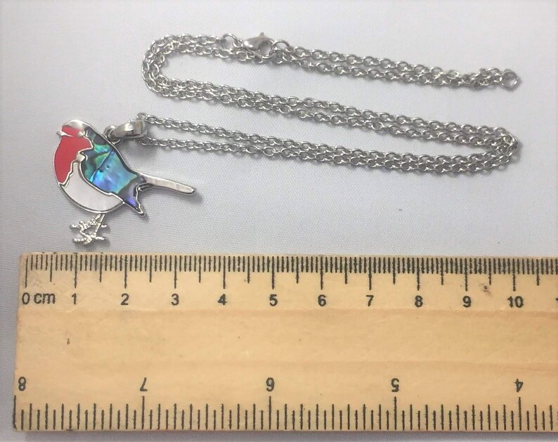 May include: A silver-colored necklace with a bird pendant. The bird pendant has a red head, white body, and iridescent blue and green accents. The necklace chain is a delicate silver color.