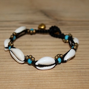 May include: A handmade bracelet with white seashells, turquoise beads, and brass details on black cord. The bracelet includes a small gold-coloured bell charm. The bracelet is designed for a comfortable fit.