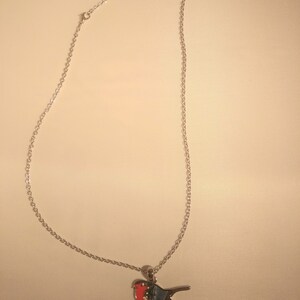May include: A silver-colored necklace with a bird pendant. The bird pendant features a red breast, blue wings, and a white belly. The necklace chain is delicate and hangs from the pendant. The bird is perched on a small, silver-colored branch.