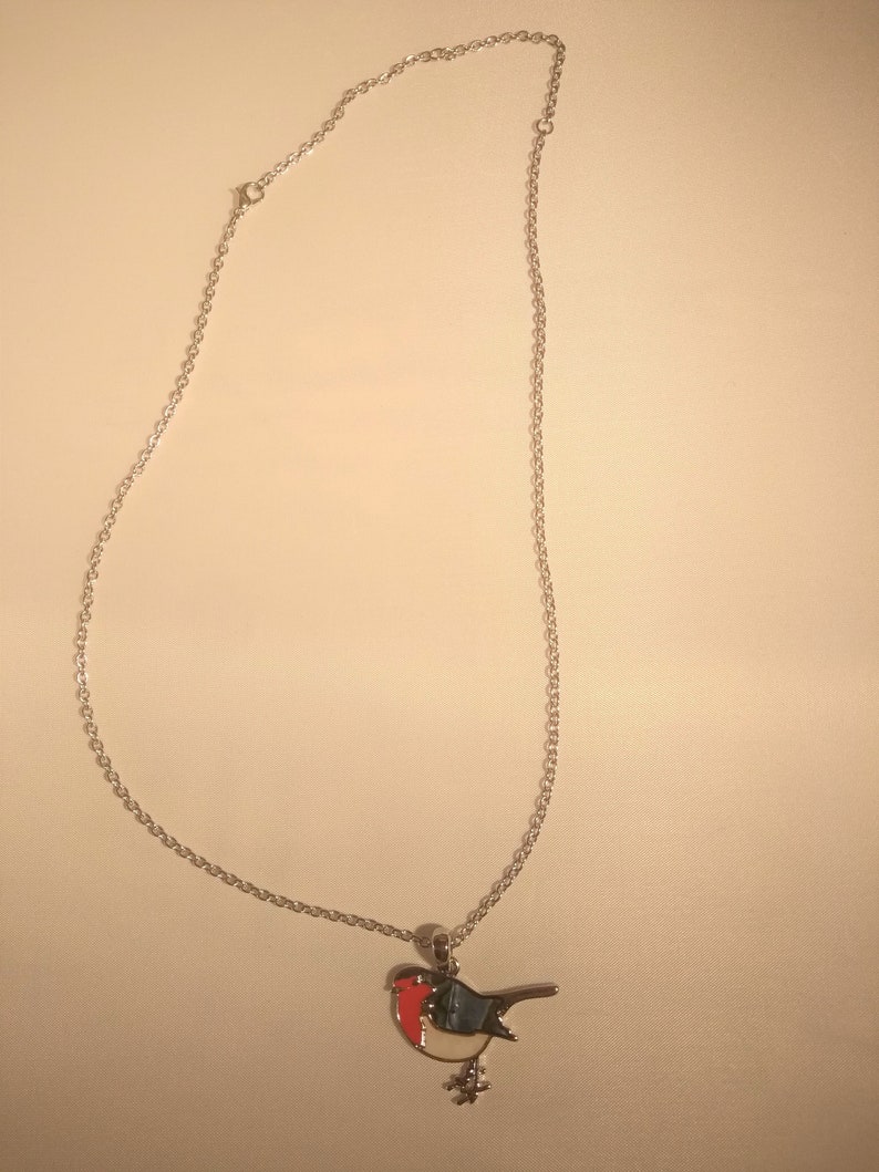 May include: A silver-colored necklace with a bird pendant. The bird pendant features a red breast, blue wings, and a white belly. The necklace chain is delicate and hangs from the pendant. The bird is perched on a small, silver-colored branch.