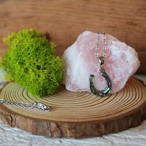 May include: A silver horseshoe pendant necklace with green and iridescent accents, displayed on a wooden slice with a pink stone and green moss. The necklace is a symbol of luck and good fortune.