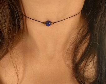 May include: A delicate choker necklace featuring a dark cord with a single, round, deep blue bead as the focal point. Small gold-colored beads flank the central bead. The necklace is worn around the neck.
