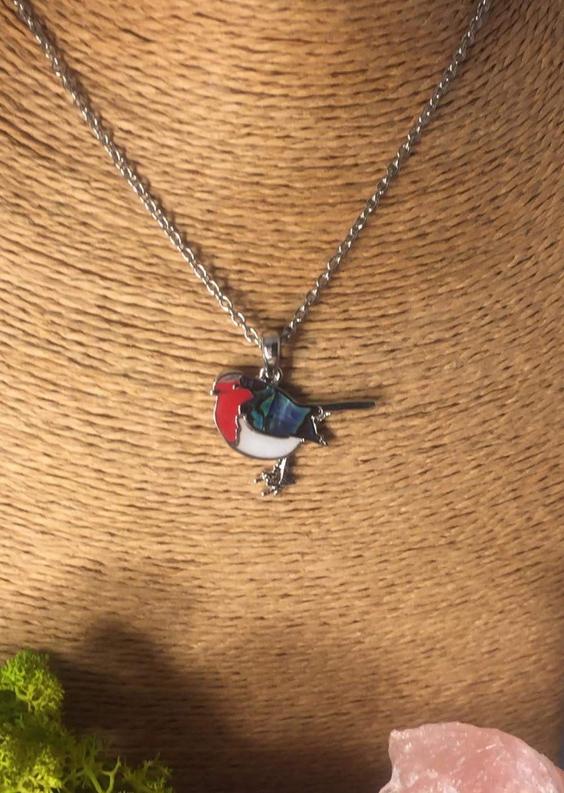 May include: A silver-toned necklace featuring a pendant in the shape of a bird. The bird pendant has a red breast, white belly, and blue and green wings. The bird is perched on a silver chain.