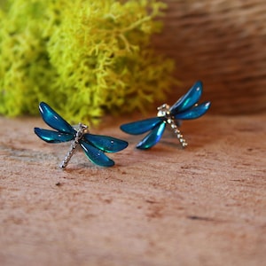 Puede incluir: Un par de pendientes de botón con forma de libélula. Los pendientes presentan alas azul verdoso con brillo metálico y cuerpos plateados. Se colocan sobre una superficie de madera con un fondo verde borroso.