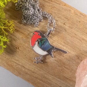 May include: A silver-toned necklace featuring a pendant shaped like a robin. The bird pendant has a red breast, white belly, and blue and green wings. The necklace is displayed on a wooden surface.