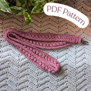 May include: A pink crocheted strap with a metal clasp. The strap is lying on a crocheted blanket with a brown and grey pattern. The text "PDF Pattern" is written in white on a pink background.