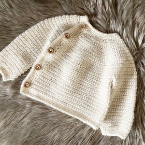 May include: Cream-colored, crocheted baby cardigan with four wooden buttons. The long-sleeved sweater is handmade with a textured stitch and is displayed on a fluffy, gray surface. A cozy and stylish garment for a baby.