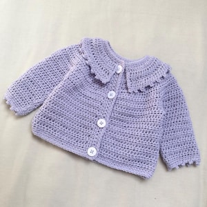 Puede incluir: Un cárdigan de ganchillo de manga larga en color violeta claro con un cuello ancho festoneado y cuatro botones blancos en la parte delantera. La prenda presenta un patrón de punto consistente en las mangas y el cuerpo, con puños festoneados.