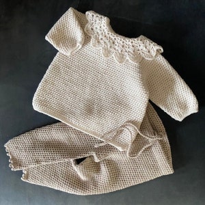 May include: A crocheted baby sweater and pants set in a light beige color. The sweater has a delicate crocheted collar with scalloped edges.