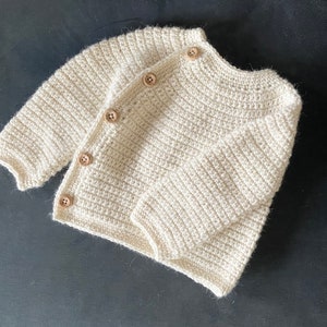 May include: Cream-colored, hand-crocheted baby cardigan with long sleeves and wooden buttons. The soft, textured knitwear is displayed on a dark surface, showcasing the detailed craftsmanship of the garment. A cozy and stylish baby clothing item.