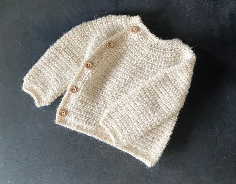 May include: Cream-colored, hand-crocheted baby cardigan with long sleeves and wooden buttons. The soft, textured knitwear is displayed on a dark surface, showcasing the detailed craftsmanship of the garment. A cozy and stylish baby clothing item.