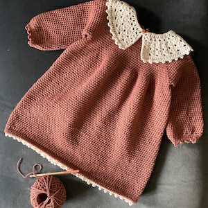 May include: A dusty rose-colored knitted dress with long sleeves and a cream-colored crocheted collar. The dress has a scalloped edge and is paired with a ball of yarn and a crochet hook, suggesting a handmade item.