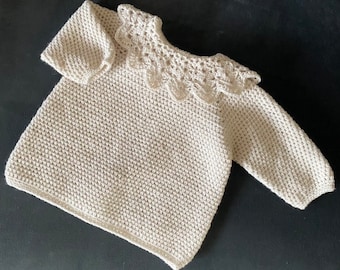 Puede incluir: Un suéter de bebé de crochet color crema, con mangas largas y cuello festoneado. El suéter está hecho de un hilo texturizado y parece suave y cómodo. El diseño es simple y clásico.