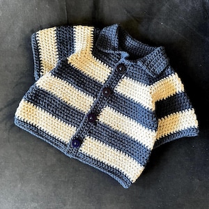 Puede incluir: Una rebeca de ganchillo de manga corta con rayas horizontales en azul marino oscuro y crema. La prenda presenta un cuello clásico, una tapeta frontal con cuatro botones circulares oscuros y un patrón de punto texturizado en todo el diseño.