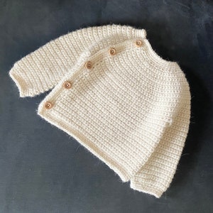 May include: Cream-colored, crocheted cardigan with long sleeves and wooden buttons. The handmade sweater is a soft, textured knit, perfect for layering. A cozy, neutral-toned garment for babies or toddlers.
