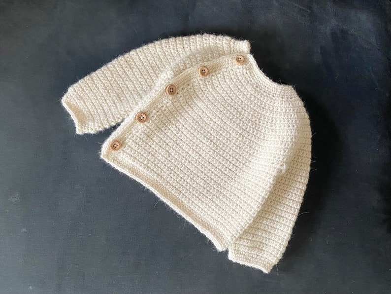May include: Cream-colored, crocheted cardigan with long sleeves and wooden buttons. The handmade sweater is a soft, textured knit, perfect for layering. A cozy, neutral-toned garment for babies or toddlers.