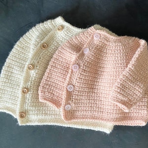 May include: Two handmade crochet baby cardigans. One is a soft cream color, the other is a light pink. Both have wooden buttons and a ribbed texture. The cardigans are displayed on a dark surface.