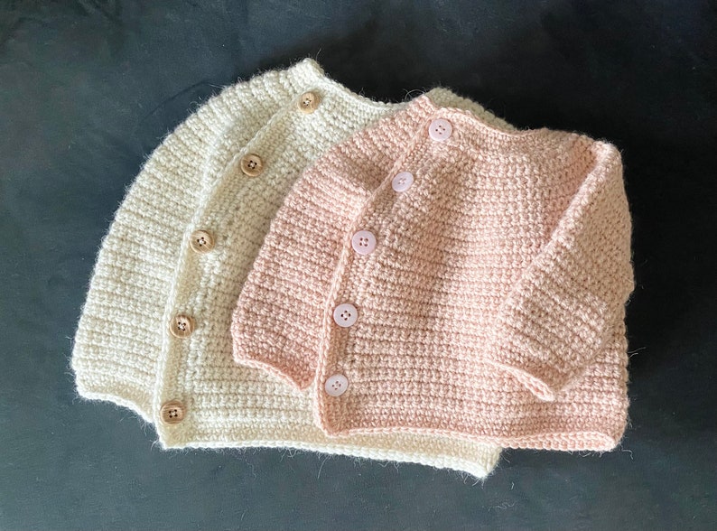 May include: Two handmade crochet baby cardigans. One is a soft cream color, the other is a light pink. Both have wooden buttons and a ribbed texture. The cardigans are displayed on a dark surface.