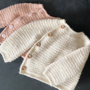 May include: Three handmade crochet baby cardigans in soft colors. The top cardigan is a light pink, the middle one is off-white, and the bottom one is a creamy white. Each cardigan has wooden buttons and a ribbed texture.